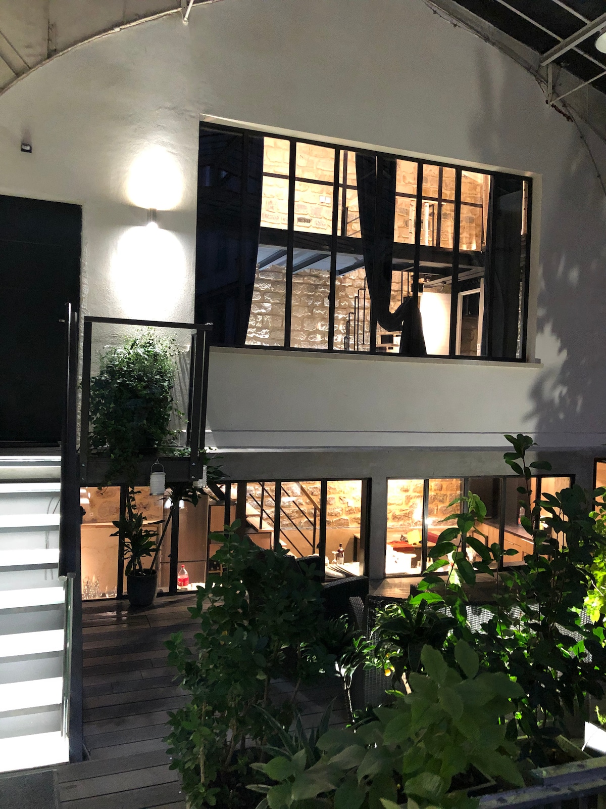 The image captures a lit-up entrance of a stylish house featuring a blend of modern and rustic architecture. Large windows reveal a glimpse of the interior's warm lighting, while lush greenery frames the outdoor space, creating an inviting ambiance.