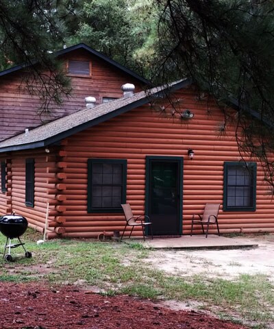 The log cabin exterior features warm wooden tones and a traditional design. Two porch chairs are positioned beside the entrance, creating a welcoming spot. A charcoal grill sits nearby, surrounded by a natural landscape of grass and trees, inviting outdoor relaxation.