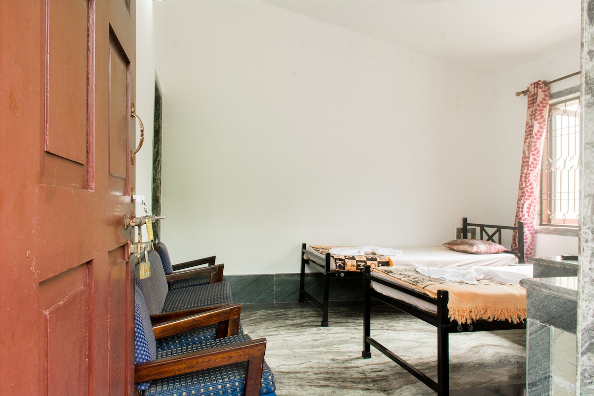 The image shows an interior room featuring two single beds and a seating area with blue chairs. A marble floor adds to the simplicity of the space, while a window draped with patterned curtains allows natural light to enter.