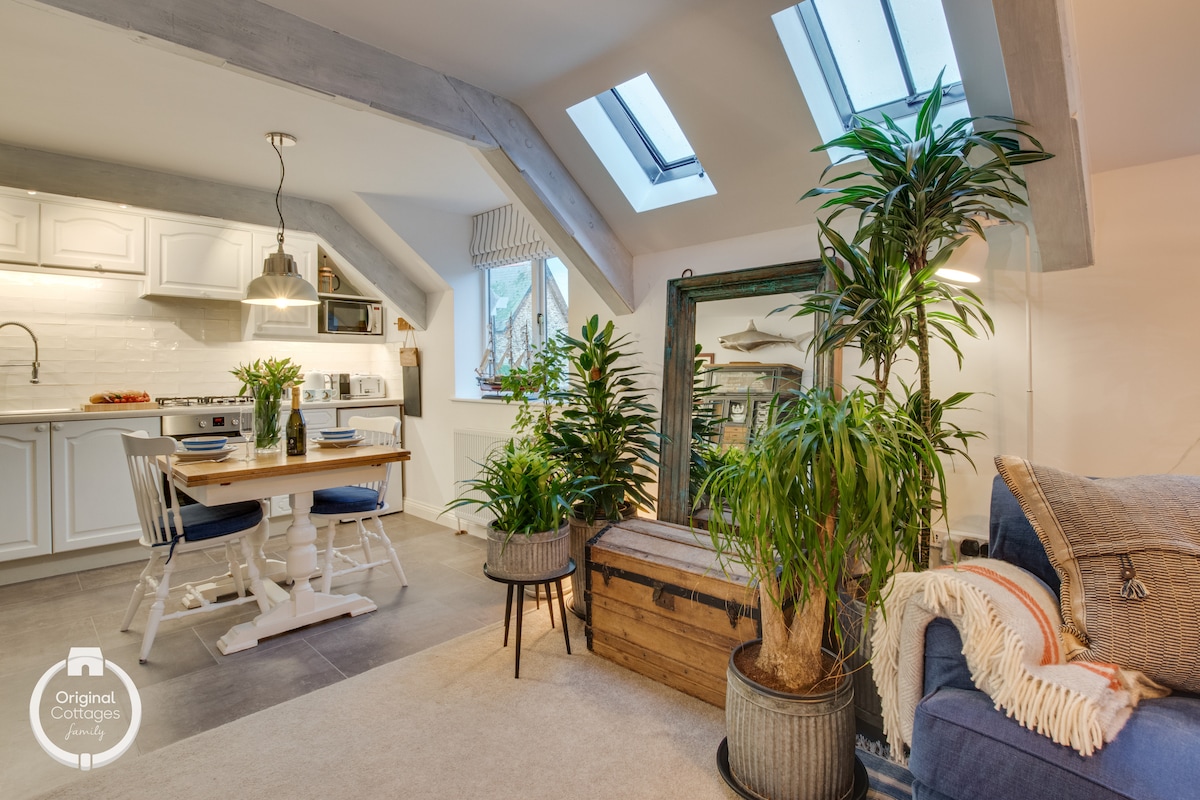 A stylish living area is complemented by large windows with skylights, allowing natural light to fill the space. A wooden dining table, surrounded by white chairs, invites meals. Lush potted plants enhance the decor, while a vintage trunk and a mirror provide functional charm.
