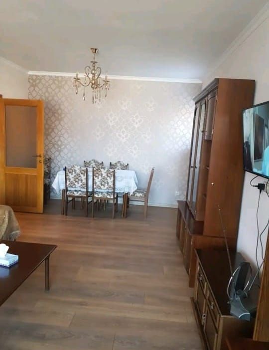 A well-lit living area showcases a dining table surrounded by six chairs, set against a patterned wall. A wooden cabinet stands against the opposite wall, while a television is mounted nearby. Warm wood tones complement the overall inviting atmosphere.