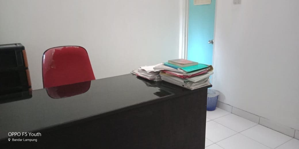 A modern office space features a sleek black desk with a red chair positioned behind it. Stacks of documents are organized on the desk, while a blue wastebasket can be seen in the corner. A light-colored door leads out of the room.