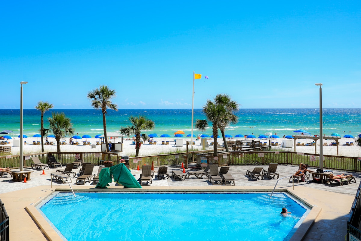A tranquil pool area is set against a backdrop of the Gulf of Mexico. Sun loungers and palm trees are positioned around the pool, which reflects the clear blue sky. Beach umbrellas dot the sandy shoreline in the distance, inviting relaxation.