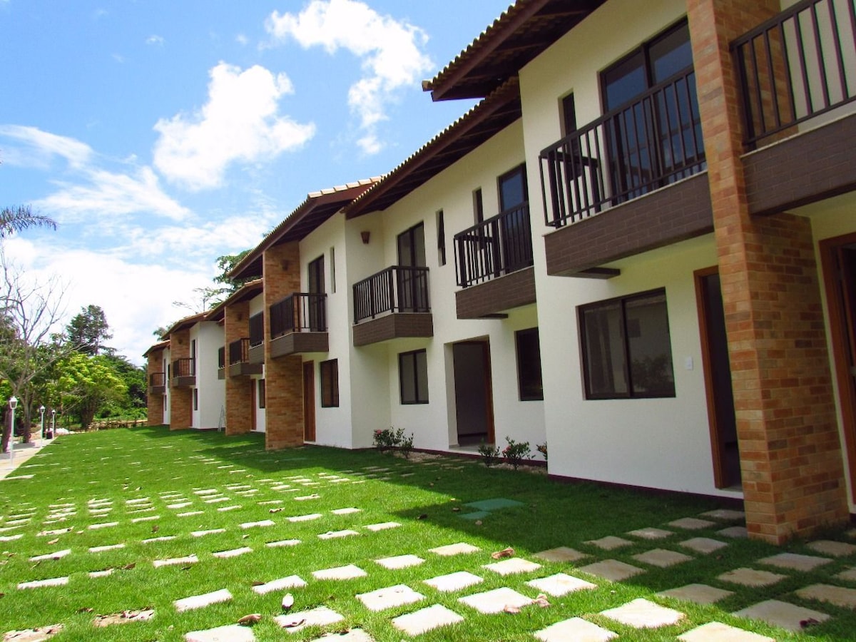 The exterior view of the two-story building showcases a row of accommodations, each featuring balconies. A well-maintained lawn with stone pathways leads to the entrance of the units, surrounded by greenery and under a clear blue sky.