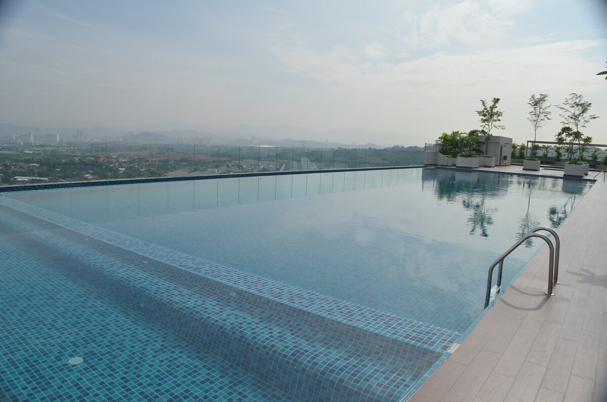An expansive infinity pool is located on a rooftop, showcasing clear blue water and a seamless view of the horizon. Surrounding planters add greenery, while the distant landscape features soft hills and a city skyline under a bright, open sky.