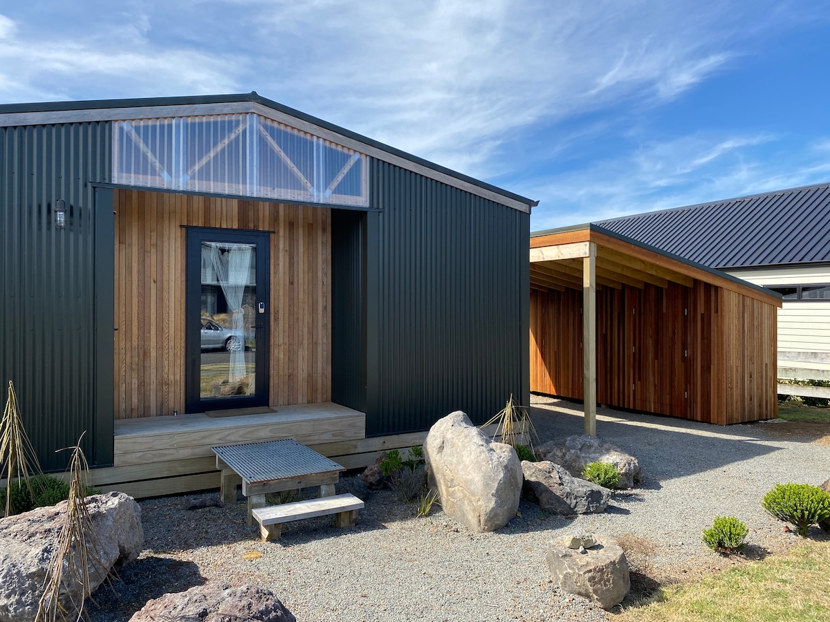 A modern cabin exterior features a wood-accented entrance with large glass doors. The surrounding landscape includes a gravel pathway, large rocks, and low-maintenance greenery. An adjacent structure offers additional covered storage space, complemented by a clear blue sky.