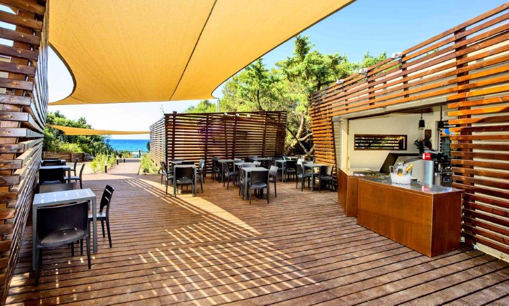 An outdoor dining area is presented, featuring wooden decking and shaded seating arrangements. Several tables with black chairs are spaced across the terrace, complemented by a wooden serving counter. The sea is visible in the background, enhancing the space's open-air feel.