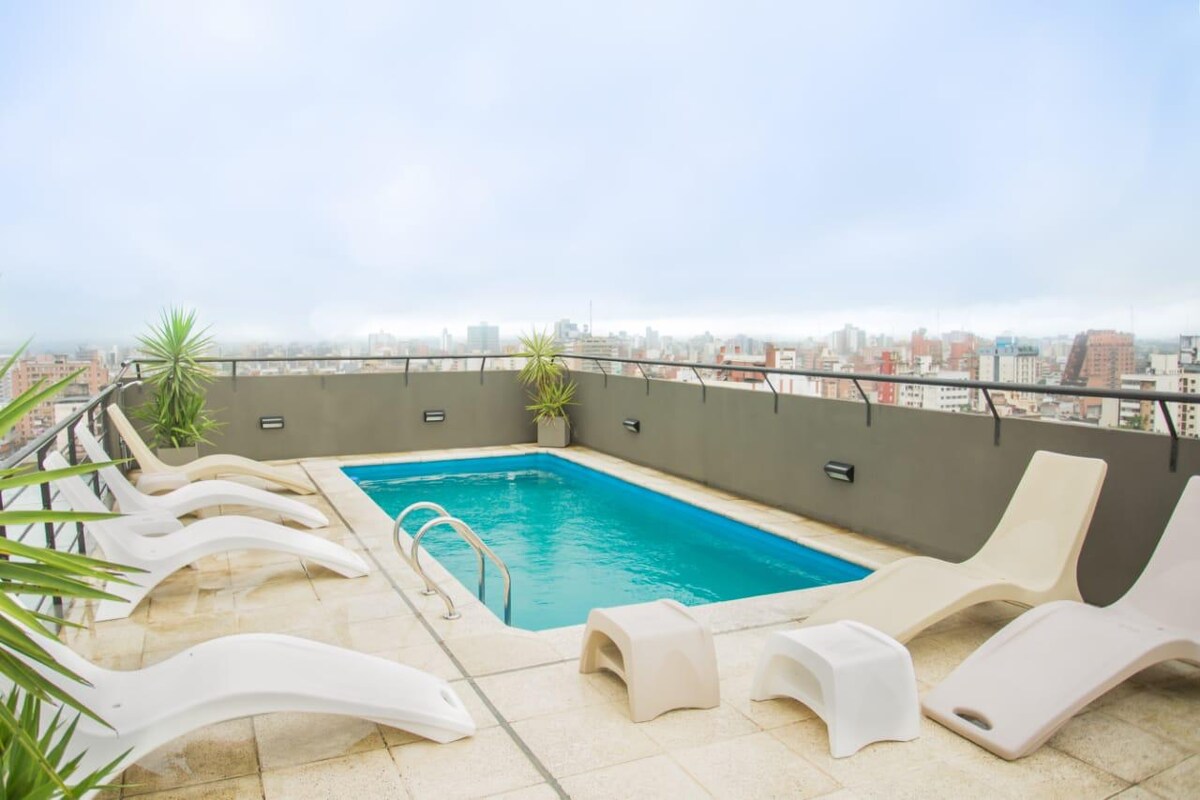 A rooftop terrace features a swimming pool surrounded by lounge chairs. Potted plants are placed along the perimeter, and a panoramic city view is visible in the background. The area is paved with light-colored tiles, enhancing the outdoor space.