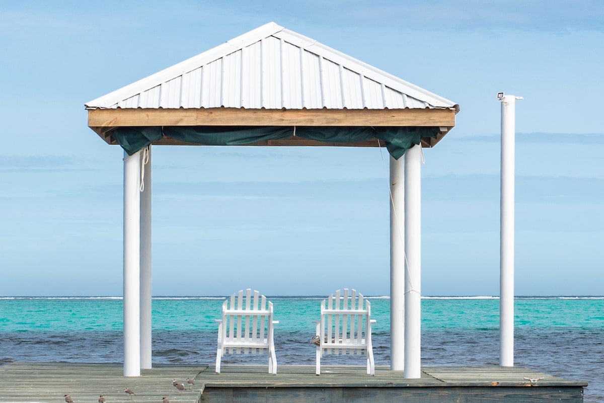 A seaside gazebo, featuring a sloped metal roof, is supported by white pillars. Two sturdy, white Adirondack chairs are positioned under the shelter, offering a view of the turquoise Caribbean Sea and the distant barrier reef.