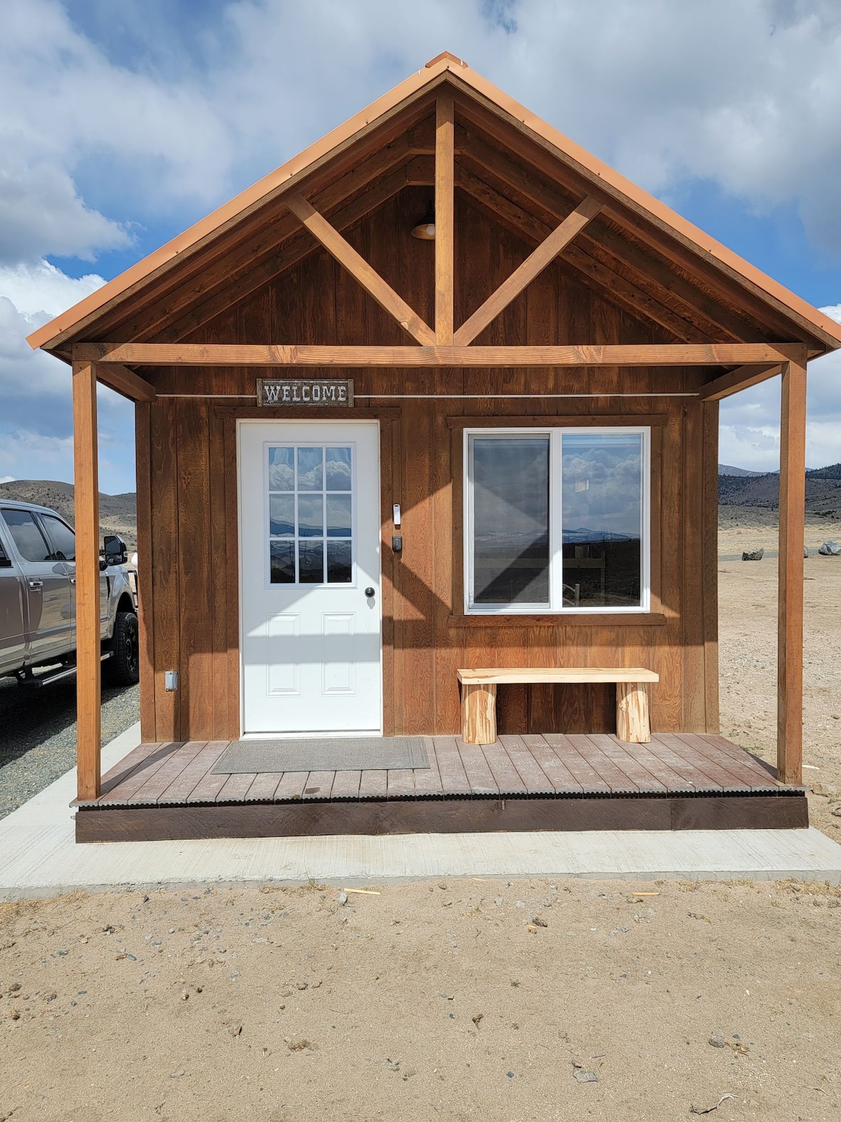 A rustic cabin exterior features wooden siding with a welcoming sign above the door. A front porch with a wooden bench provides a cozy entryway. A large window allows natural light in, framing distant mountain views against a partly cloudy sky.