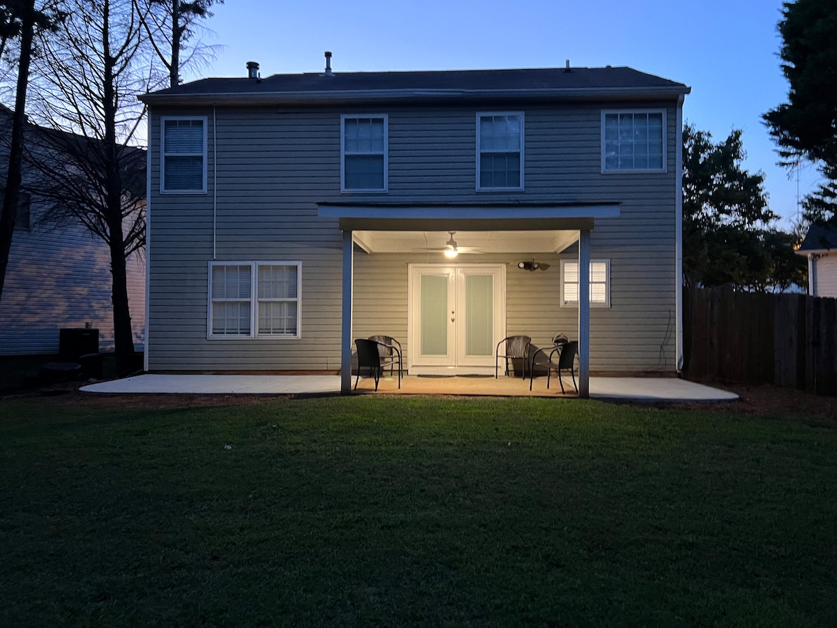 A two-story house is shown with a covered porch featuring two chairs and a table. Light is emanating from the porch area, illuminating the surroundings. The backyard is spacious, with well-maintained grass and a wooden fence visible in the background.