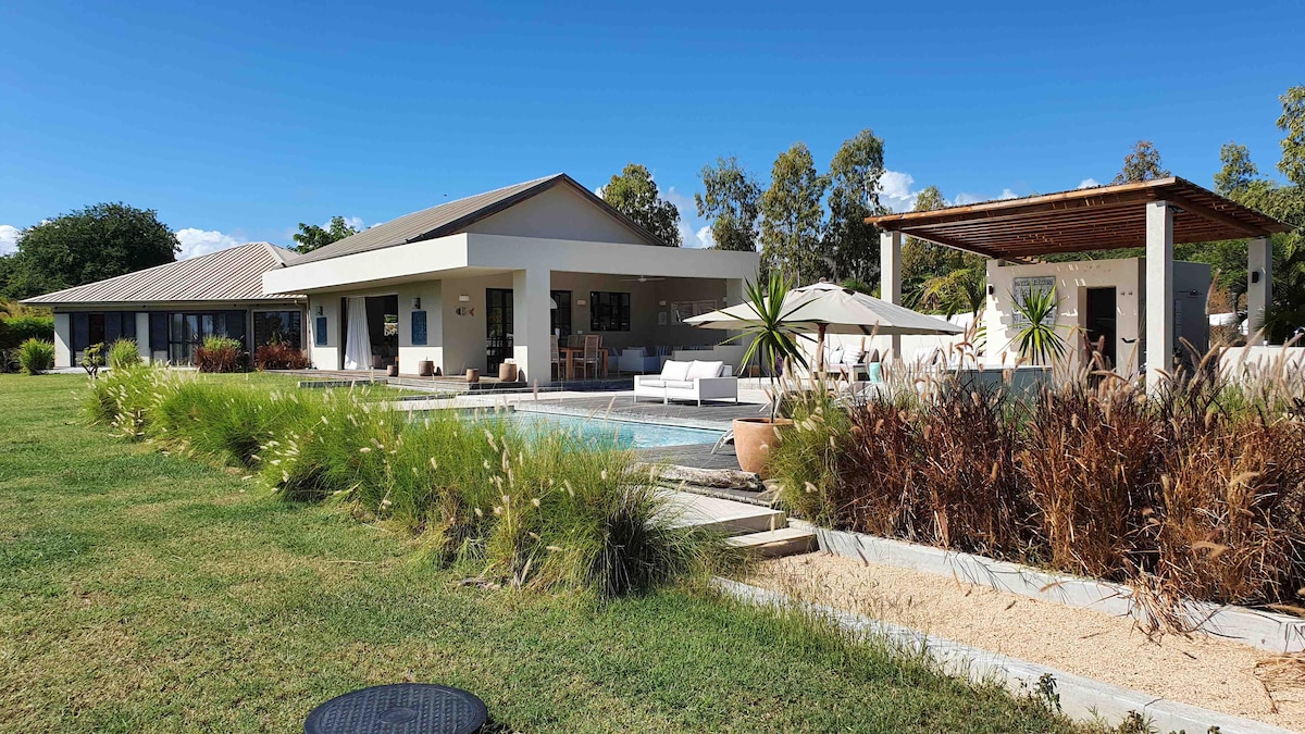 A modern villa is set amidst lush greenery, featuring a large outdoor pool and a shaded terrace. The architecture includes a spacious main building with extensive glass elements and a separate structure for relaxation. Sun loungers and tropical plants enhance the outdoor living area.