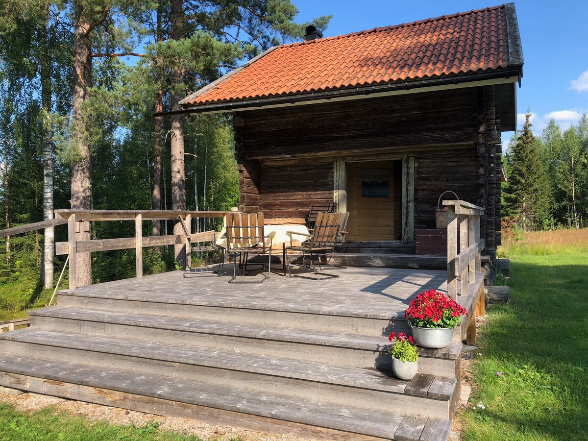 A charming wooden building stands surrounded by lush greenery, featuring a red-tiled roof. A spacious wooden deck is equipped with outdoor seating and flower pots, providing a comfortable area to enjoy nature. The entrance door is clearly visible, blending with the rustic design.
