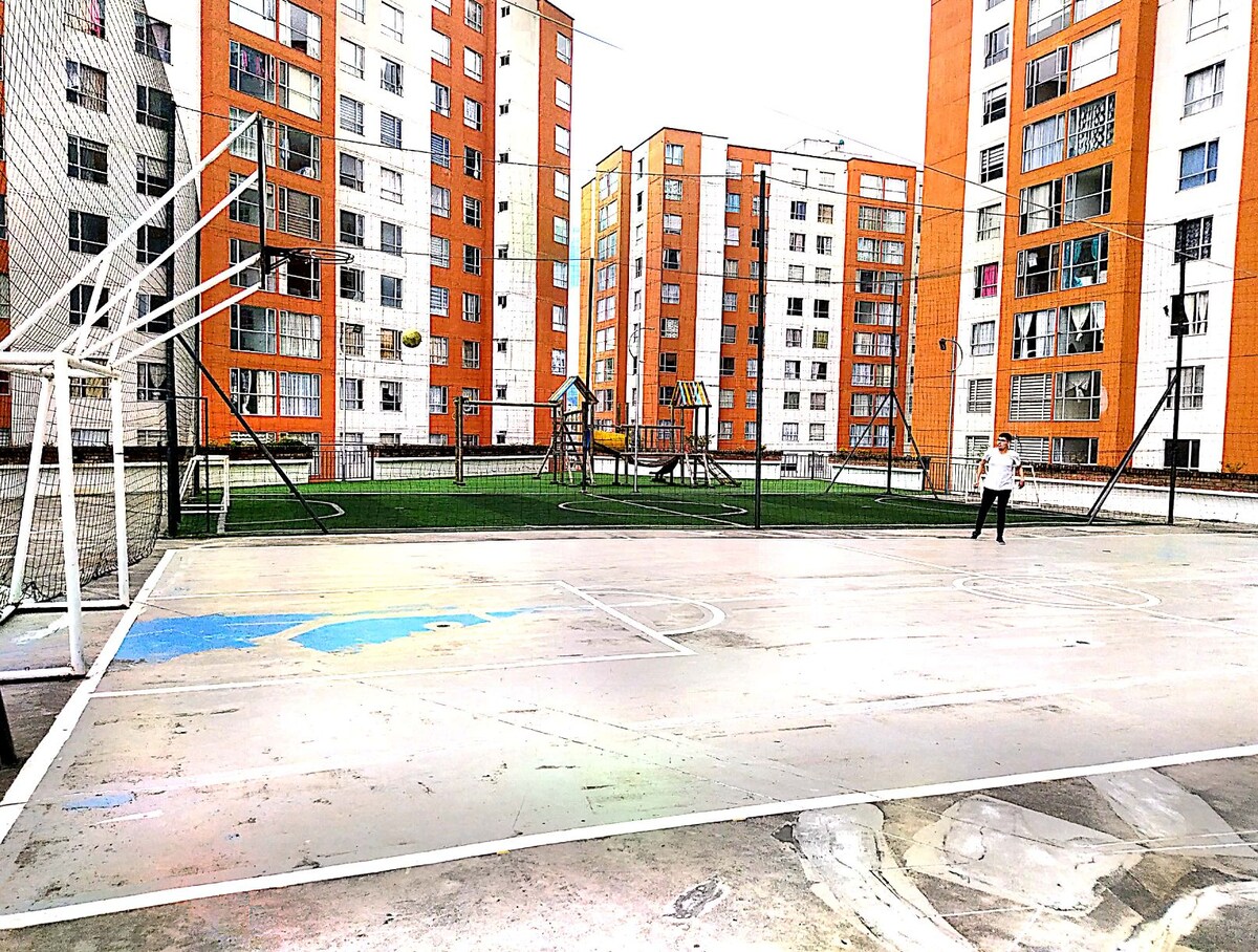A multi-sport playing area is visible, featuring a basketball court marked with faded lines. Surrounding it are several residential buildings with orange and white facades. A playground with climbing equipment is positioned on the grassy field beyond the court.
