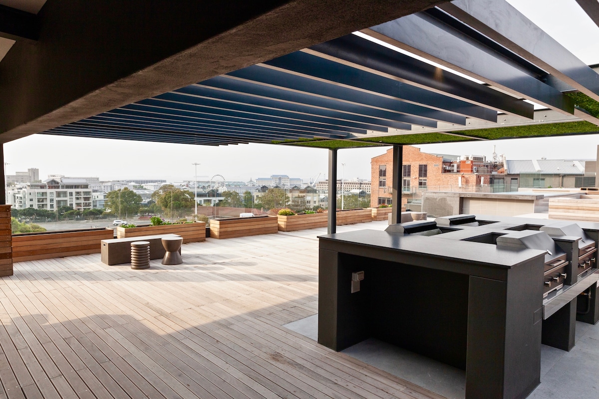 A spacious rooftop deck is shown, featuring an outdoor kitchen area with modern appliances. Comfortable seating arrangements are visible alongside wooden planter boxes. A panoramic city view is framed by the sleek covering above, enhancing the open-air atmosphere.