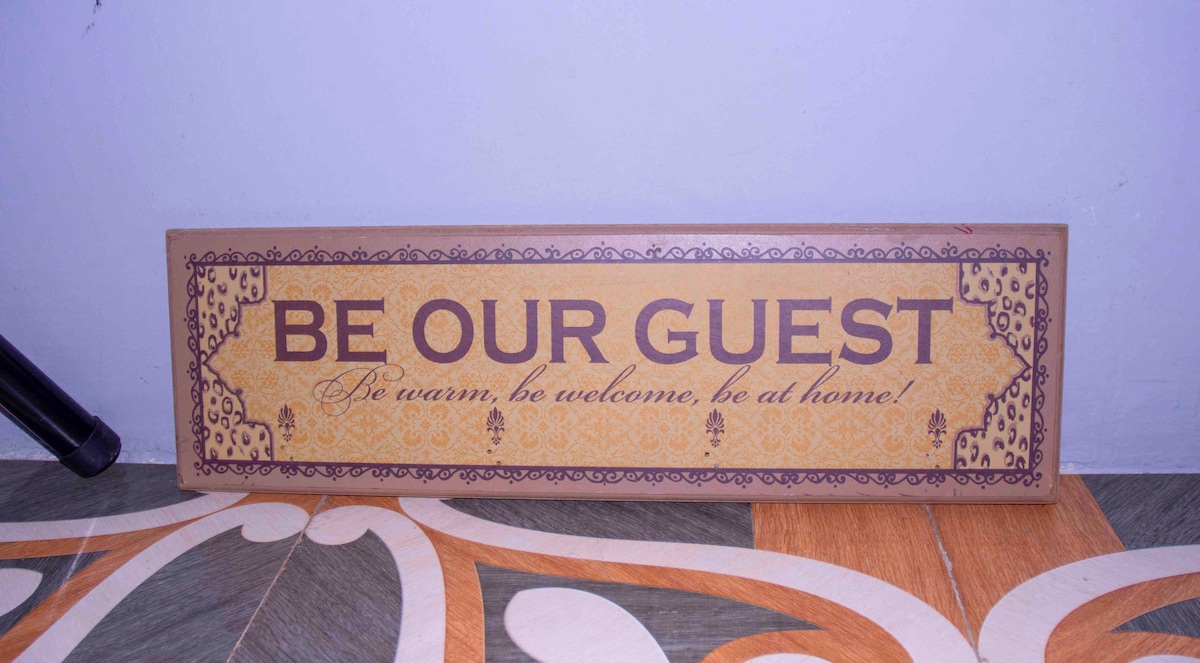 A decorative wooden sign featuring the phrase 'Be Our Guest' in bold letters. The sign is framed with intricate designs and warm colors, resting on a patterned floor that incorporates earthy tones. The inviting sentiment is clearly conveyed.