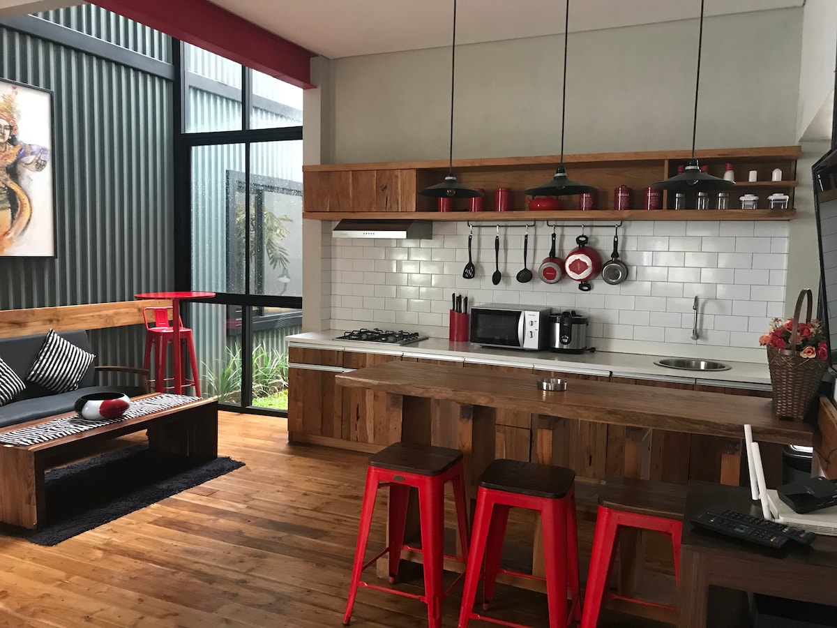 An open kitchen area features wooden cabinetry and modern appliances. Red bar stools accompany a wooden dining counter. A large window allows natural light to illuminate the space, complemented by greenery visible outside. Art decor adds a touch of character to the walls.