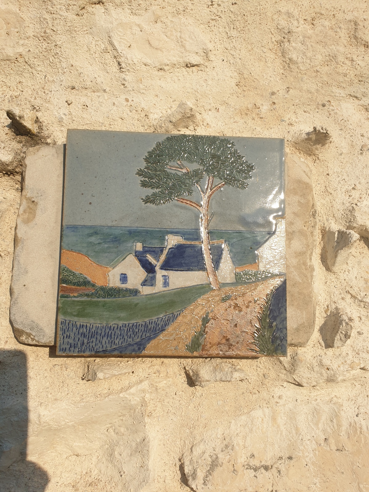A ceramic tile art piece is affixed to a stone wall, depicting a simplified landscape with a cottage and a tree. A winding pathway leads towards the cottage, while fields and water are shown in the background under a clear sky.