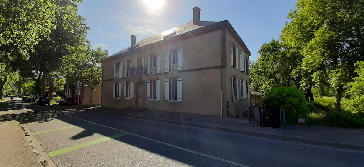 A historic village house is positioned along a tree-lined street. The two-story structure features traditional architecture with numerous shuttered windows, bordered by well-maintained greenery. Sunlight bathes the facade, contributing to a serene atmosphere.