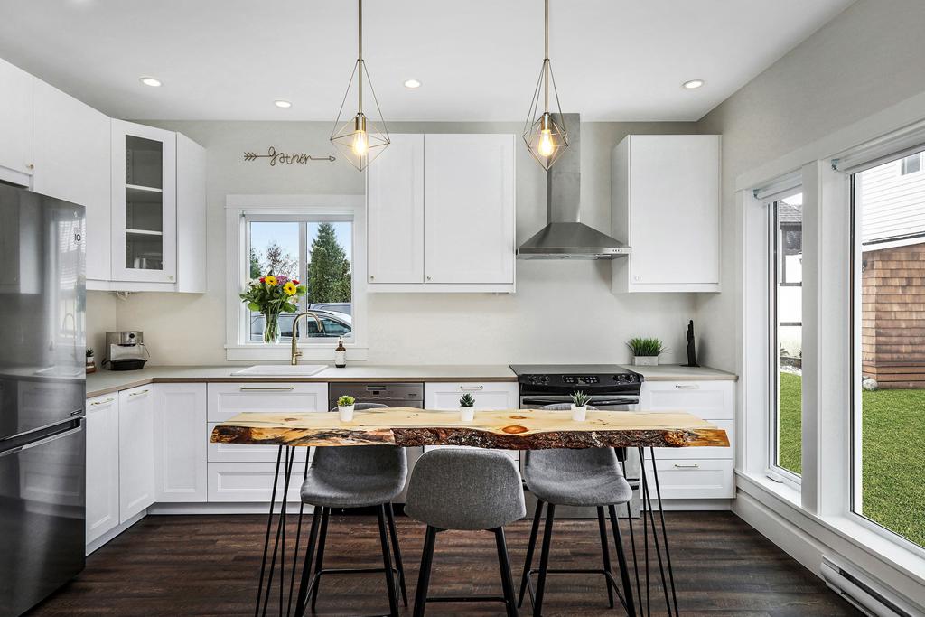 A modern kitchen features sleek white cabinetry and stainless steel appliances. A wooden dining table with four grey chairs is centered under two pendant lights. Natural light is provided by large windows overlooking an outdoor space, enhancing the bright and clean atmosphere.