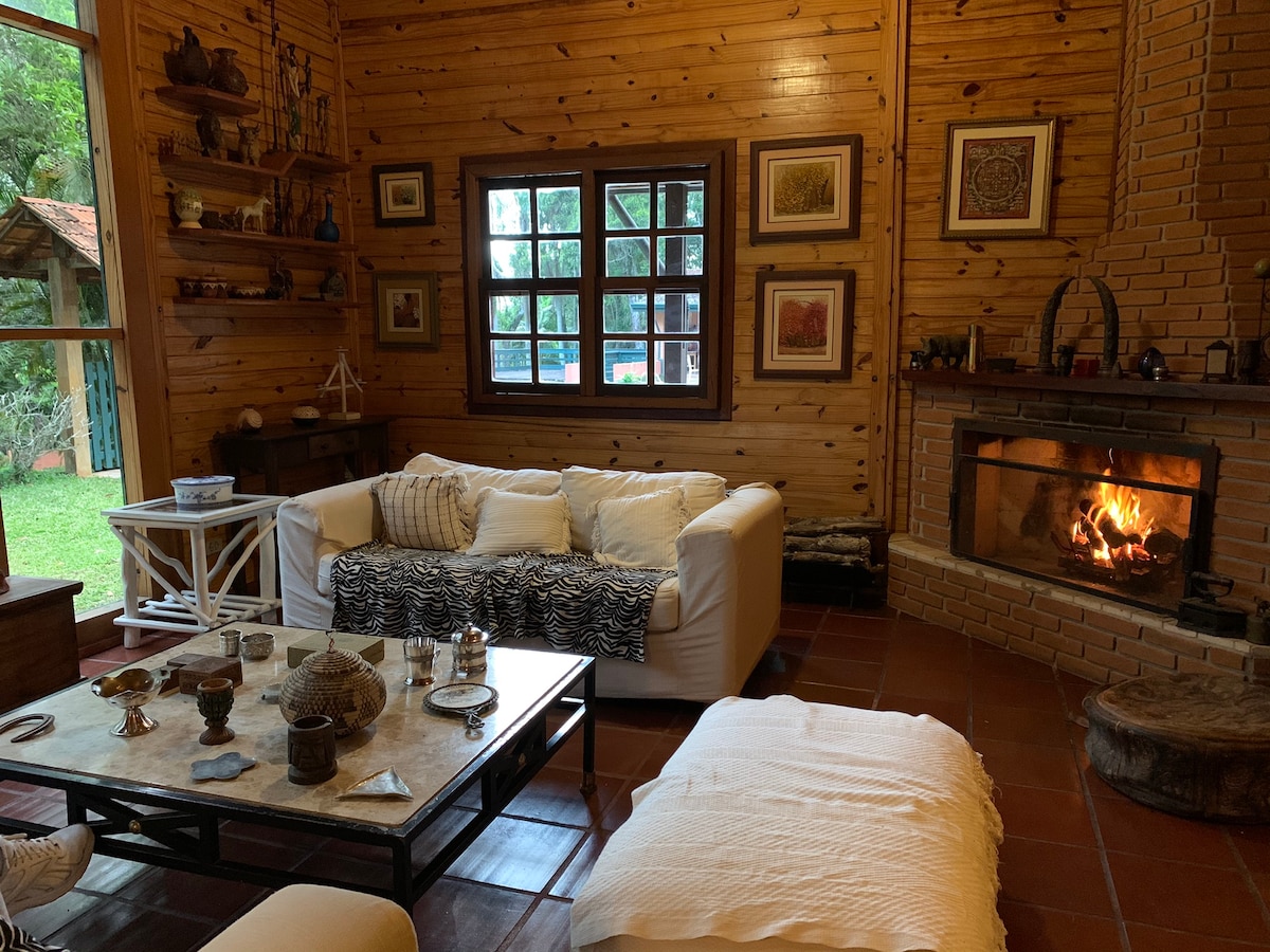 A cozy living room features wooden walls and a large fireplace. A white sofa with a striped blanket is paired with a coffee table adorned with decorative items. Light enters through a window, enhancing the warm ambiance of the space.