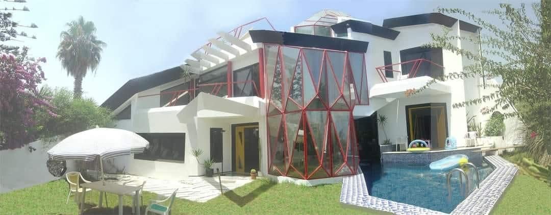 The villa features a modern architectural design with a distinct red geometric frame. A spacious terrace extends towards the swimming pool, surrounded by well-maintained gardens. Sun loungers and an umbrella are positioned for relaxation, while a glimpse of seating areas is visible on the patio.