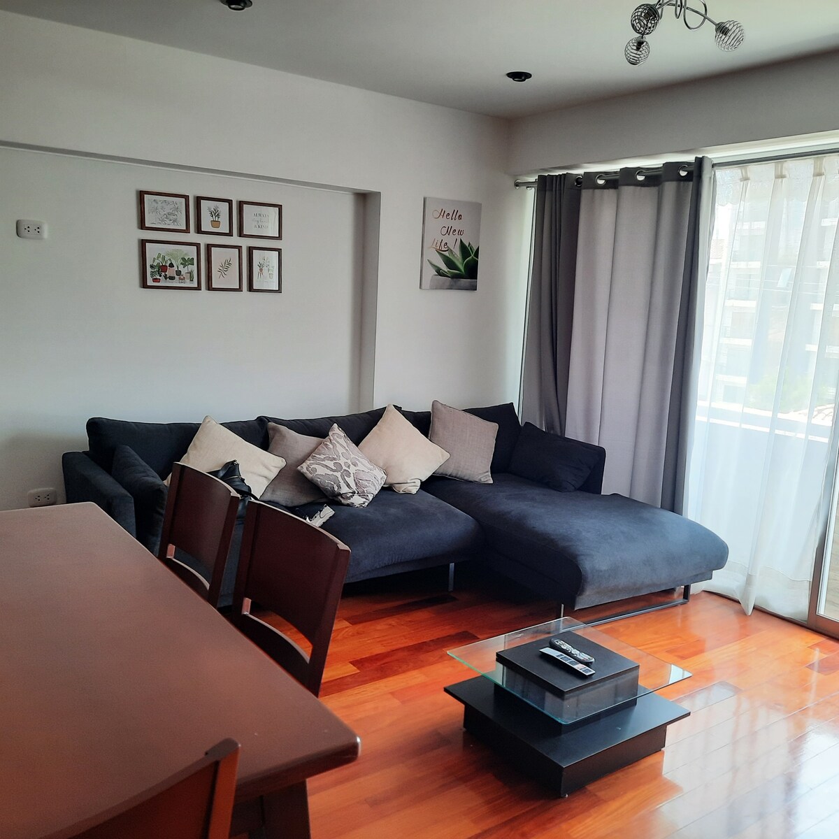 A spacious living area features a comfortable sectional sofa adorned with several decorative pillows. A sleek glass coffee table sits centrally, while a dining table with wooden chairs is positioned nearby. Natural light floods the room through large windows covered with soft drapes.