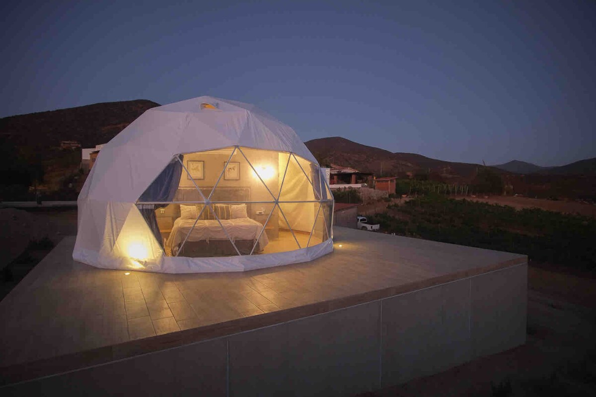 A geodesic dome stands illuminated against the night sky, showcasing a spacious bed visible through the panoramic clear window. The dome is situated on a raised platform, surrounded by rolling hills and vineyards, blending contemporary design with natural surroundings.