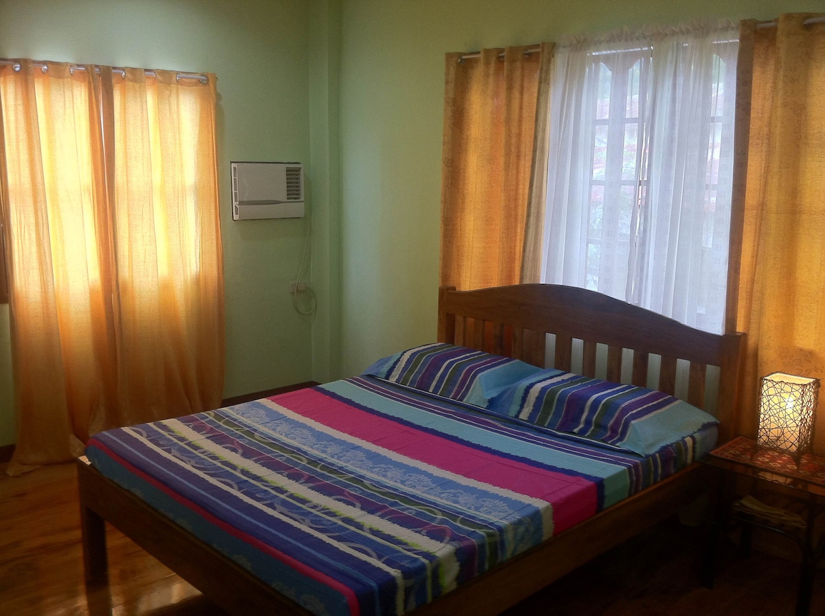A sleeping area features a spacious bed with colorful striped bedding. Soft natural light filters through sheer white curtains, complemented by warm orange drapes. An air conditioning unit is positioned on the wall, and a small lamp adds illumination beside the bed.