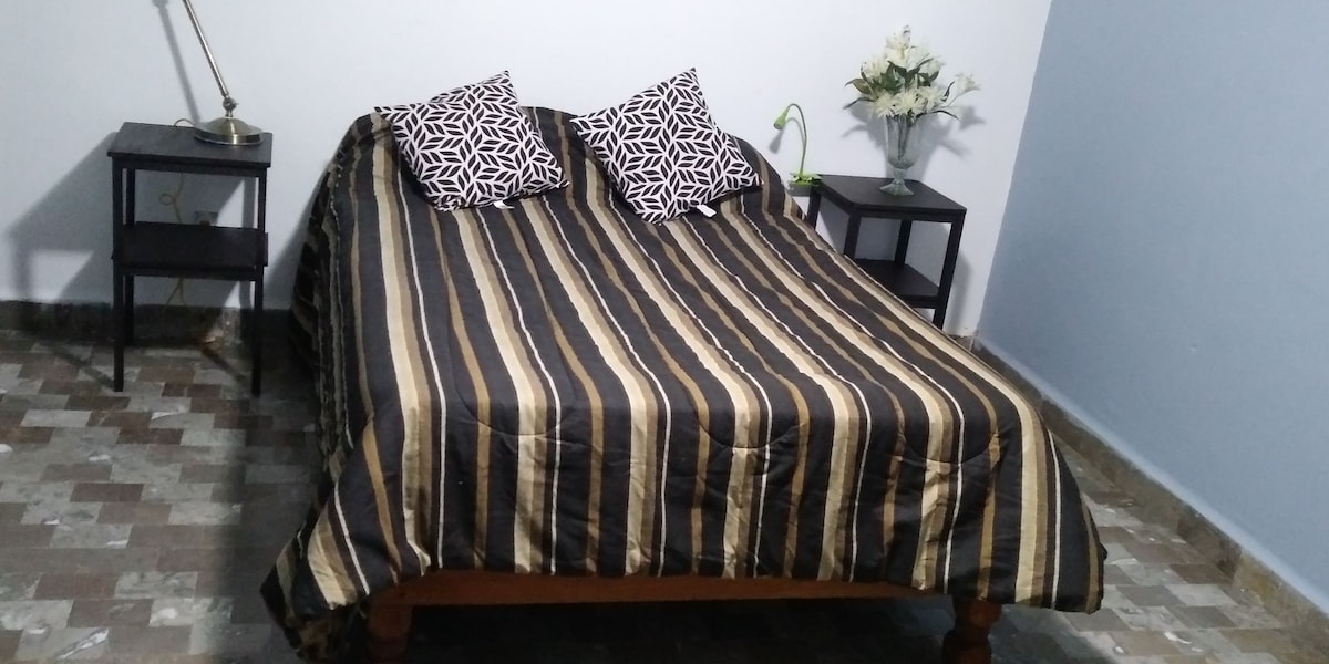 A spacious bed is covered with a striped comforter, featuring two patterned pillows at the head. On either side of the bed, there are two nightstands. A small vase with flowers is placed on one nightstand, while the floor displays a patterned tile design.