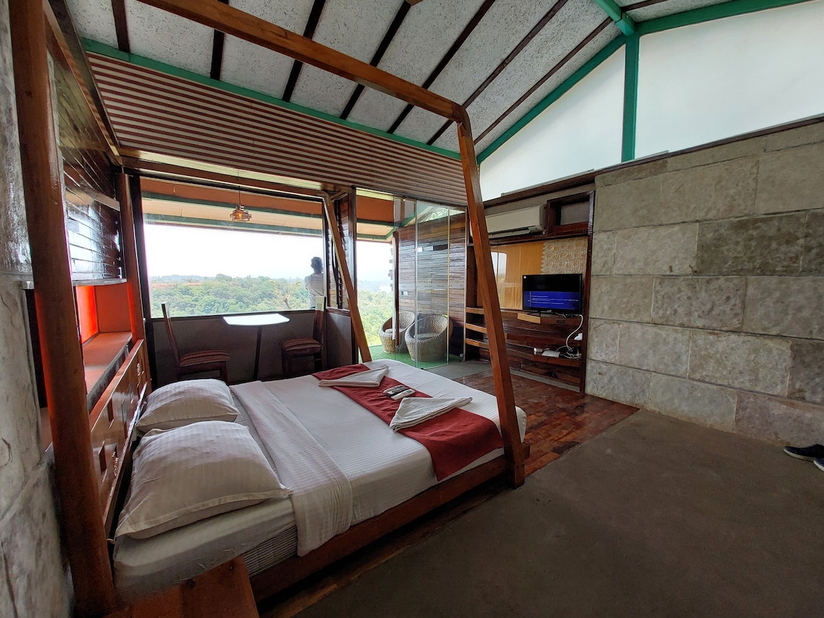 A spacious room features a large bed adorned with white linens and a vibrant red throw. Large windows provide expansive views of the surrounding landscape. A flat-screen TV and wooden furniture complete the inviting living area, creating a calm and relaxing environment.