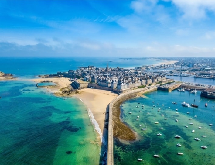 An aerial view captures the historic walled city of St. Malo surrounded by serene waters. Sandy beaches and a vibrant coastline are visible, with boats gently anchored in the harbor. The clear blue sky enhances the coastal landscape, offering a glimpse of the region's natural beauty.
