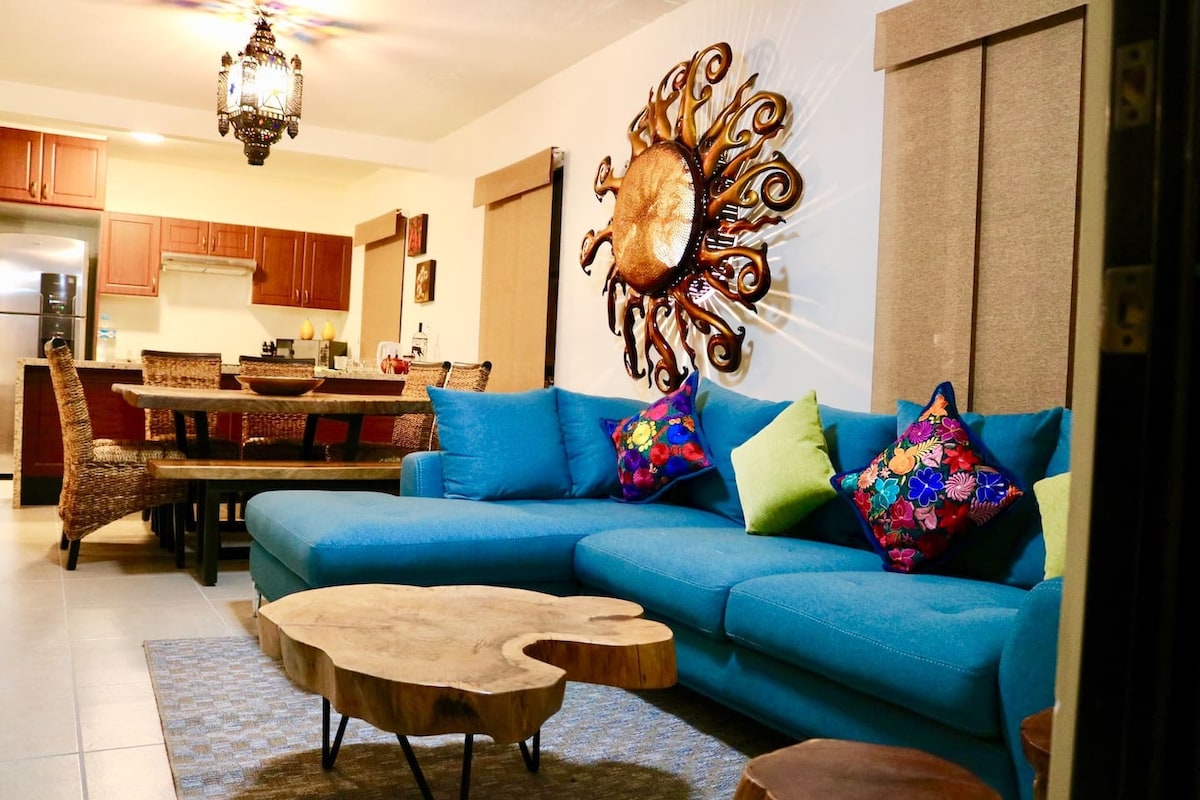 A comfortable living area is highlighted by a large blue sectional sofa adorned with colorful throw pillows. The space features a round wooden coffee table and a dining table visible in the background. Accents include wall art and warm lighting from a decorative ceiling fixture.
