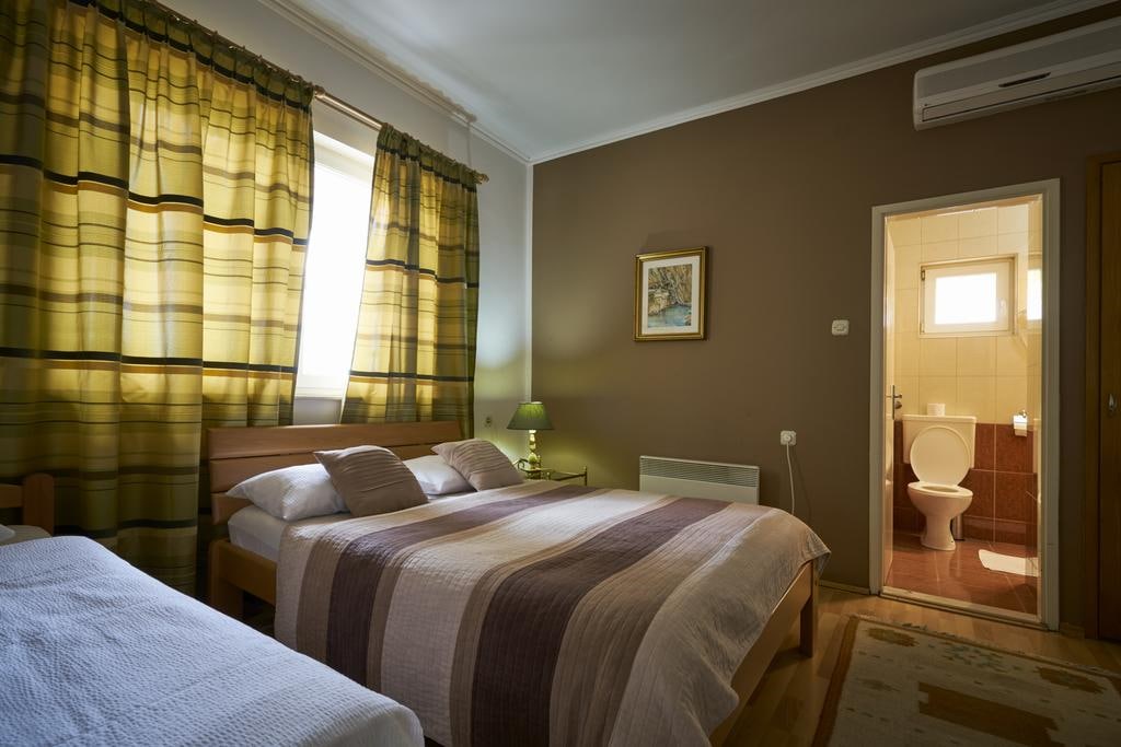 A cozy room features a spacious bed covered in neutral-toned linens, complemented by a bedside lamp on a wooden nightstand. Sunlight filters through patterned curtains, while a private bathroom is visible in the background, showcasing tiled walls and essential fixtures.