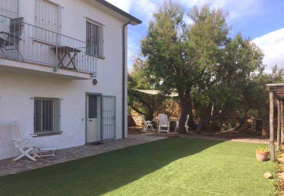 The exterior of the property features a well-maintained garden area with artificial grass. A cozy seating area is located beneath a mature tree, providing shade. The building showcases a pair of balcony doors on the upper floor, while a door leads to a ground-level entrance.