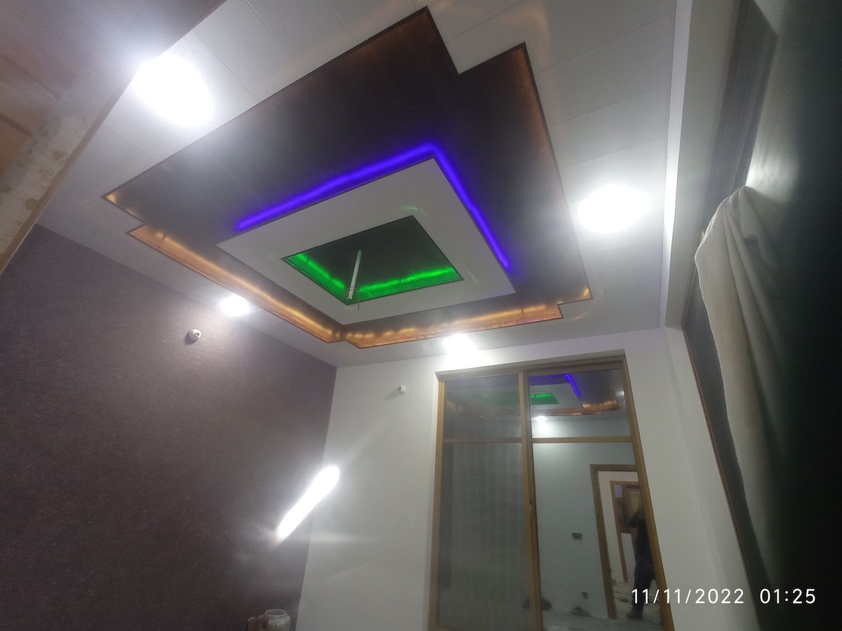 A modern ceiling design features LED lighting elements in green and blue. Recessed lights are arranged thoughtfully around the perimeter, illuminating the space. A large window allows natural light to enter, complementing the contemporary decor of the room.