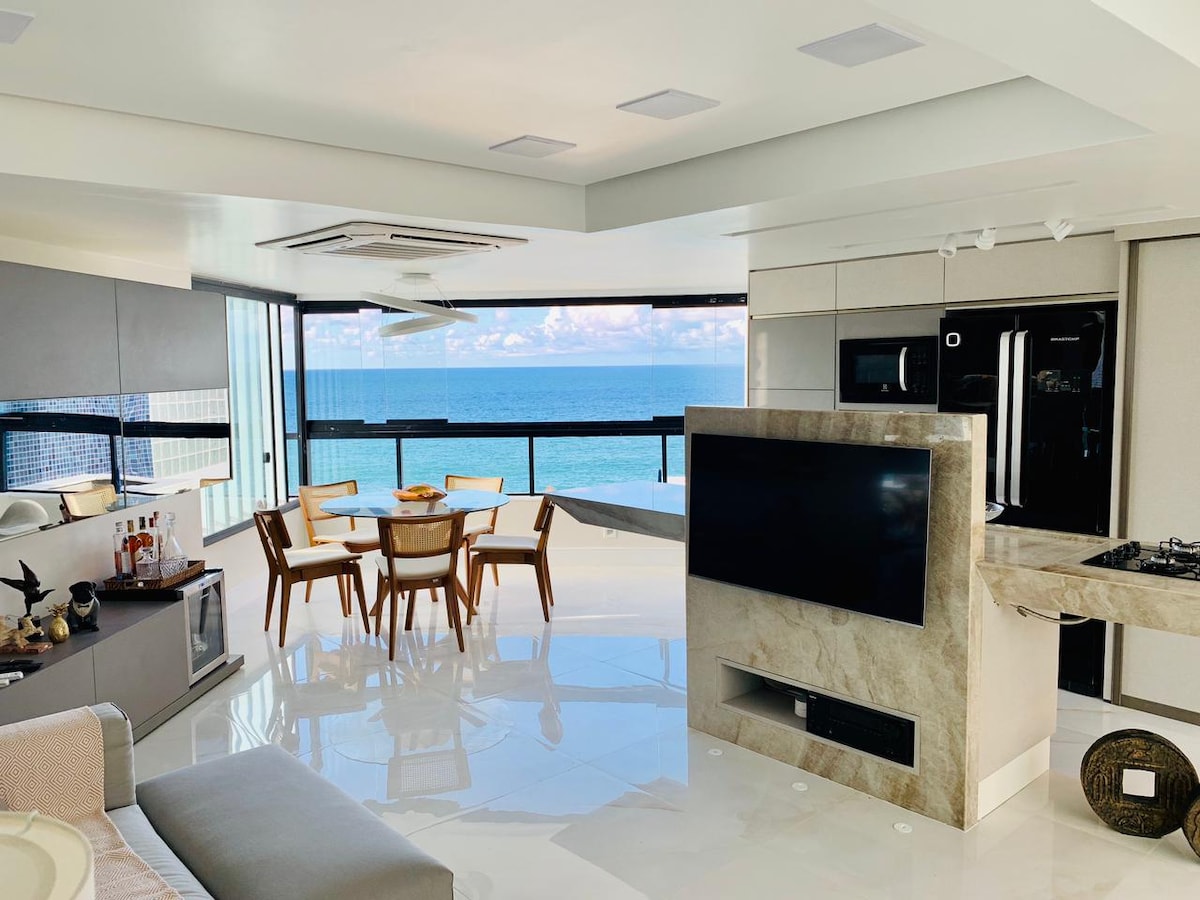 A modern living space features a sleek kitchen and dining area, with a circular table surrounded by six chairs. Floor-to-ceiling windows provide a striking view of the ocean, and light reflects off the glossy floor, enhancing the open and airy feel.