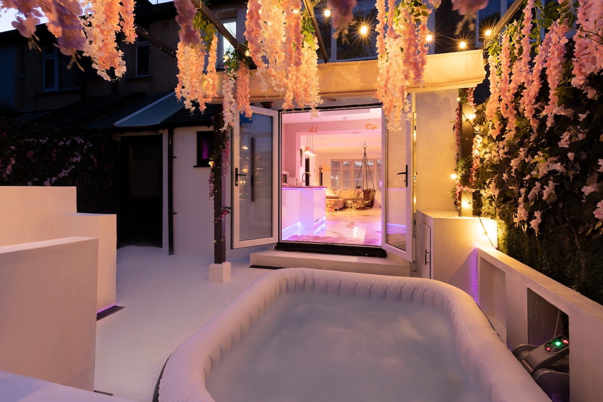 An outdoor area is highlighted by a white hot tub surrounded by soft lighting and floral decorations. Open doors provide a view into a tastefully decorated indoor space. The ambiance is complemented by warm tones and hanging lights, creating an inviting atmosphere.