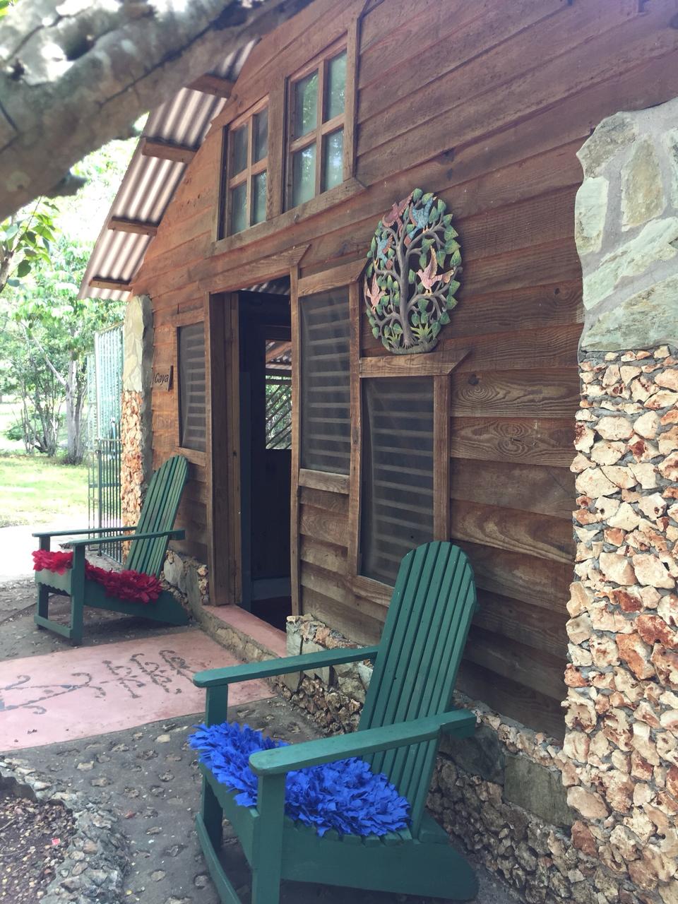 A charming exterior of a wooden cabin is shown, featuring a façade made of timber and stone. Two green adirondack chairs are positioned on a pathway, adorned with red and blue decorative items. The entrance is framed by windows with wooden shutters, inviting a sense of warmth.