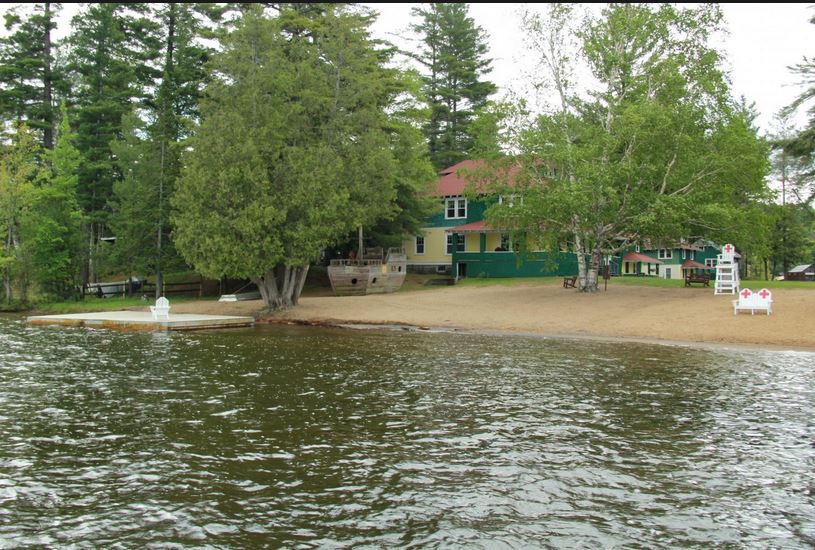 A sandy beach extends along the edge of a serene lake, bordered by tall trees. A grassy area is visible with a wooden deck and steps leading to the water. A charming two-story house in the background features green siding, complementing the natural surroundings.