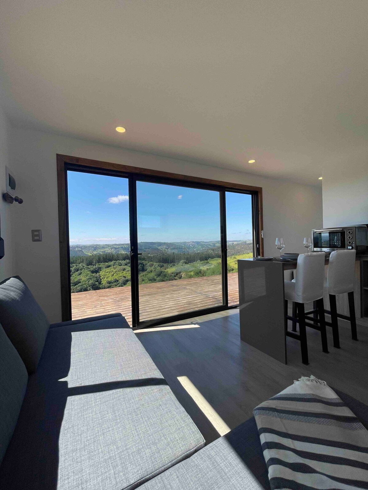 A modern living space is featured with large glass doors that open to a panoramic view of the valley. Natural light floods the area, highlighting the sleek kitchen and dining setup. A cozy sofa is positioned nearby, creating an inviting atmosphere to enjoy the scenery.