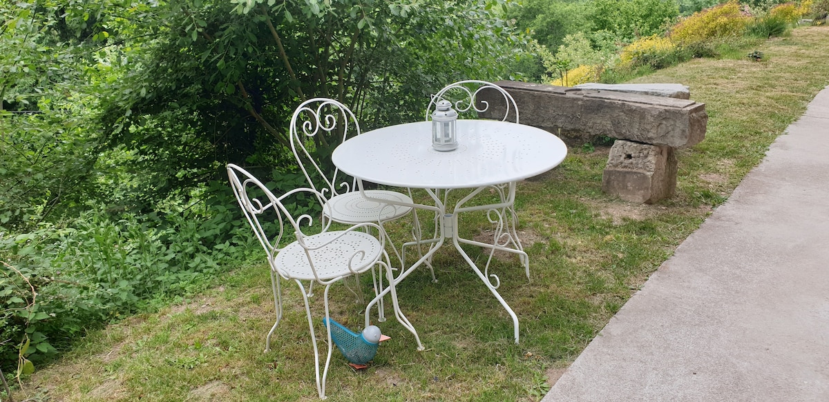 An outdoor seating area is featured, consisting of a round white metal table and two matching chairs. The table is adorned with a lantern, and a decorative bird can be seen nearby. Lush greenery and wildflowers create a natural backdrop.