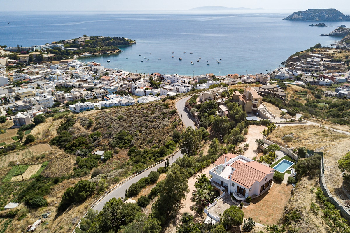 An aerial view captures a serene villa surrounded by lush greenery, with a private pool and expansive outdoor area. The coastline of Agia Pelagia is visible in the background, dotted with boats and framed by charming village architecture, highlighting the villa's proximity to the beach.