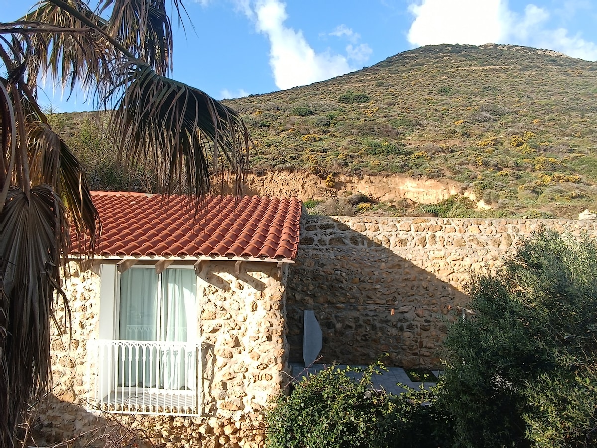 A stone structure is shown with a terracotta-tiled roof. Light filters through a window with a balcony, framed by lush greens. A rocky hill rises in the background, partially covered with vegetation, offering a natural and serene setting.