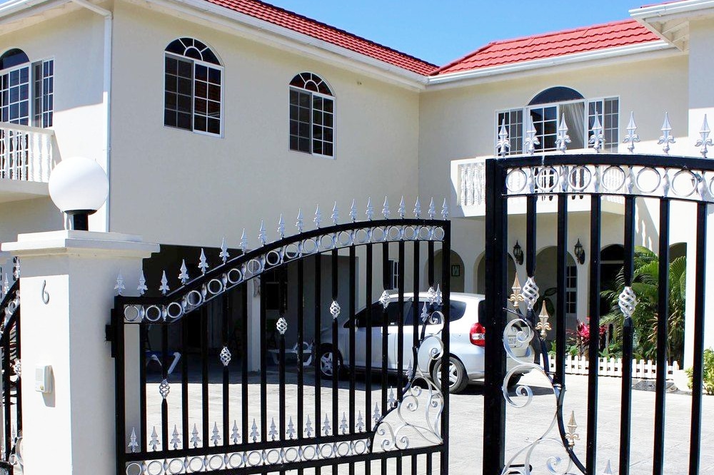 An elegant landscaped entrance features a decorative black iron gate that opens to a spacious driveway. A clean white vehicle is parked, while the two-story structure showcases large windows with arched tops and a red tiled roof, creating a welcoming facade.