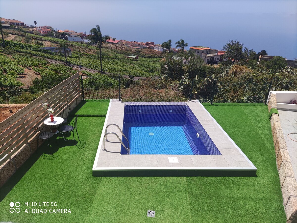 An inviting infinity pool is set against a backdrop of rolling vineyards and distant coastal views. The pool area features sleek tiles and a ladder for easy access. A small table with two chairs is positioned nearby on a grassy surface, ideal for relaxation.