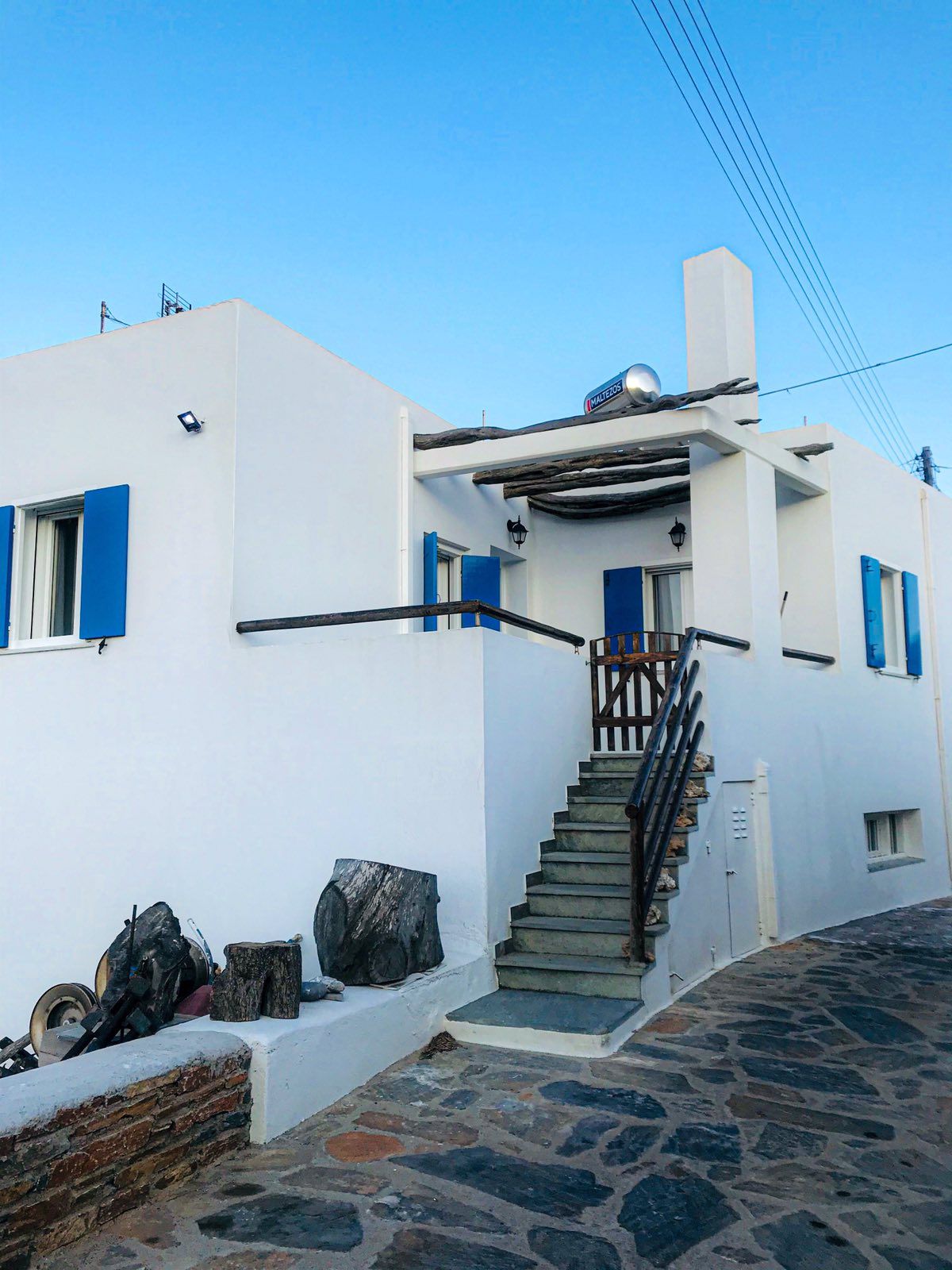 A newly constructed Cycladic house features a clean white façade and contrasting blue shutters. Stairs ascend to the entrance, framed by a rustic wooden railing. Natural stone pathways lead up to the property, highlighting a serene village atmosphere.
