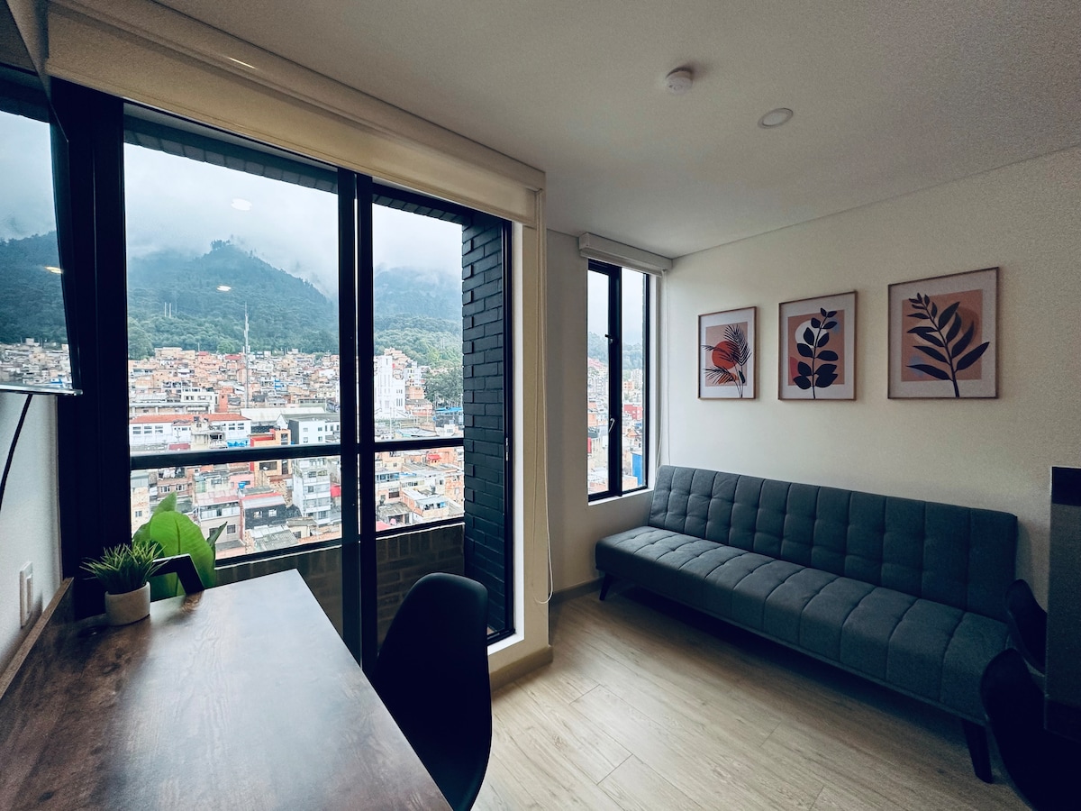 A modern living space is showcased, featuring a gray sofa and a wooden desk with a chair. Large windows offer a panoramic view of the city and mountains beyond. Three art prints adorn the wall, adding subtle decorative elements.