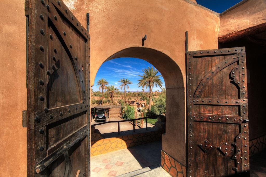 A grand entrance features intricately designed wooden doors, ajar to reveal a view of palm trees swaying gently in the breeze. The sky overhead is clear with a hint of blue, creating a welcoming pathway towards the inviting surroundings outside.