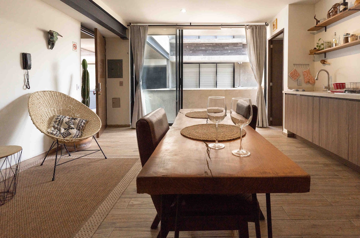 A wooden dining table is set for two with glassware, placed on a textured rug. A modern wicker chair is visible beside the large glass doors, which open to a balcony, allowing natural light to fill the spacious interior.
